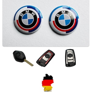 Może przedstawiać: Dwa okrągłe, białe, niebieskie, czerwone i czarne emblematy samochodowe BMW. Trzy kluczyki samochodowe z czarnymi, czerwonymi i srebrnymi brelokami. Mapa Niemiec z kolorami niemieckiej flagi.