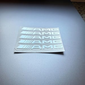 Könnte beinhalten: Ein Bogen mit fünf hellblauen AMG-Logo-Aufklebern. Jeder Aufkleber zeigt das AMG-Logo in weißer, stilisierter Schrift mit einem parallelen Liniendesign auf der linken Seite. Die Aufkleber befinden sich auf einer transparenten Folie.