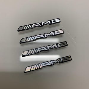 May include: Four AMG emblems. Each emblem is rectangular with the letters "AMG" in white, and a white striped design on the left side. The emblems have a silver and black colour scheme.