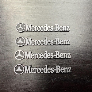 May include: Four silver Mercedes-Benz emblems are displayed on a dark surface. Each emblem features the Mercedes-Benz logo, a three-pointed star within a circle, and the brand name in a sans-serif font.