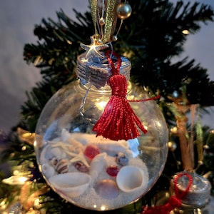 May include: A clear glass Christmas ornament filled with white sand and seashells. The ornament has a red tassel hanging from the top and is decorated with a gold star.