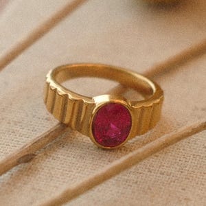 May include: A gold ring featuring a ruby-coloured oval gemstone. The ring has a textured band and is displayed with yellow floral accents on a neutral background. Jewellery.