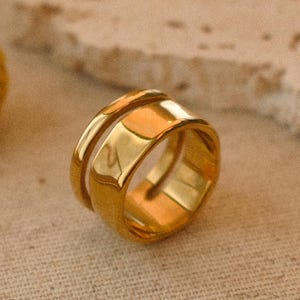 May include: A gold ring featuring a double-band design. The ring is made of a shiny, reflective metal and rests on a neutral-toned surface. The image's focus is the ring, with yellow floral accents in the background.