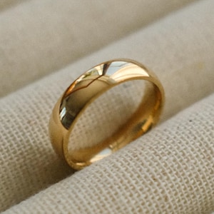 Può includere: Un anello dorato con una superficie liscia e lucida. L'anello è posto su uno sfondo di tessuto beige chiaro. L'anello sembra essere una fede nuziale o un tipo di gioiello simile.