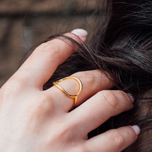 May include: A gold ring featuring a teardrop design. The ring is worn on a hand, with dark hair in the background. The teardrop shape of the ring is open in the middle.
