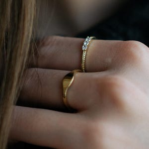 Peut inclure: Une bague chevalière en or à la surface lisse et polie, accompagnée d'une seconde bague en or dotée d'un anneau fin serti de trois petites pierres claires et scintillantes. Les deux bagues sont portées aux doigts, avec une finition métallique.