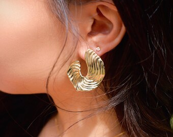 May include: Gold hoop earrings with a wavy, textured design. The earrings are worn by a person with long black hair.