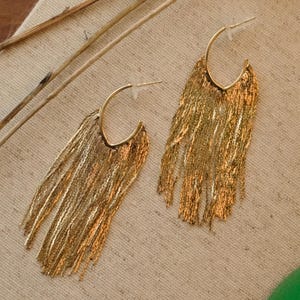 May include: A pair of gold-colored hoop earrings with a fringe design. The earrings are made of thin, delicate strands that create a flowing, textured effect. The earrings are displayed on a neutral-colored fabric.