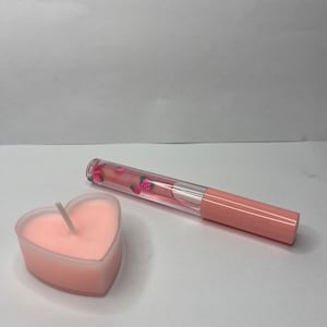 May include: A pink heart-shaped candle with a white wick and a pink lip gloss tube with a clear, strawberry-filled body.