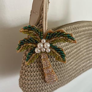 Peut inclure: Un sac à main beige tissé avec un charm en forme de palmier perlé. Le charm en forme de palmier présente des perles vertes et dorées pour les feuilles, des perles nacrées pour les noix de coco et un tronc perlé beige. Le sac à main a une sangle en cuir beige.
