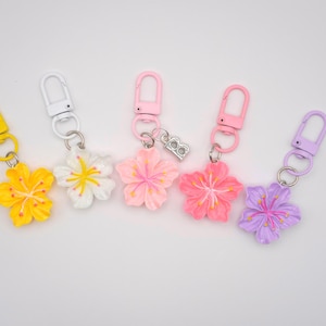May include: Five flower-shaped keyrings in yellow, white, pink, and purple. Each keyring has a different colour flower with a matching clasp. One pink keyring has a silver charm with the letter "B".