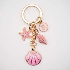 May include: A gold-coloured keyring featuring pink seashell charms. The charms include a starfish, a seashell, a clam shell, and a circular charm with the letter "R". The keyring is a beach-themed accessory.