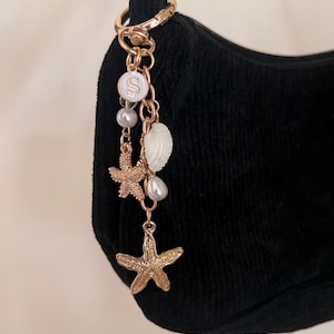 May include: A gold-colored charm with starfish, pearls, and a seashell hangs from a black handbag. The charm includes a white circular pendant with the letter "S" in the center. The handbag is made of a black, corduroy-like material.
