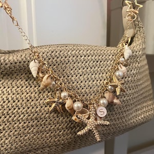 May include: A beige woven handbag with a gold chain and seashell charms. The charms include starfish, pearls, and various seashells. A gold star-shaped clasp is attached to the bag.