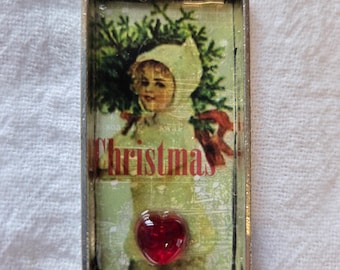 May include: Rectangular pendant with a silver-tone frame. The pendant features a vintage image of a child in a winter outfit holding a Christmas tree branch, with the word "Christmas" in red. A red heart-shaped charm is at the bottom.