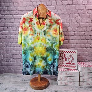 May include: A short-sleeved, button-down shirt with a vibrant tie-dye pattern in shades of red, yellow, green, and blue. The shirt is displayed on a wooden mannequin. A stack of white boxes with red text and a cartoon figure are visible.