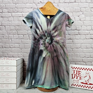 May include: A tie-dye dress with a V-neck and short sleeves, featuring a spiral pattern in shades of gray, purple, and green. The dress is hanging on a wooden hanger. The image also shows a box with the text "TIE DYE Fresh New Jersey".