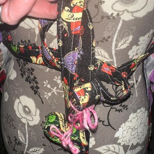 May include: A black fabric heart-shaped ornament with a pink yarn tie. The ornament is decorated with colorful tattoo-style designs and the word "Due". The background is a floral pattern.