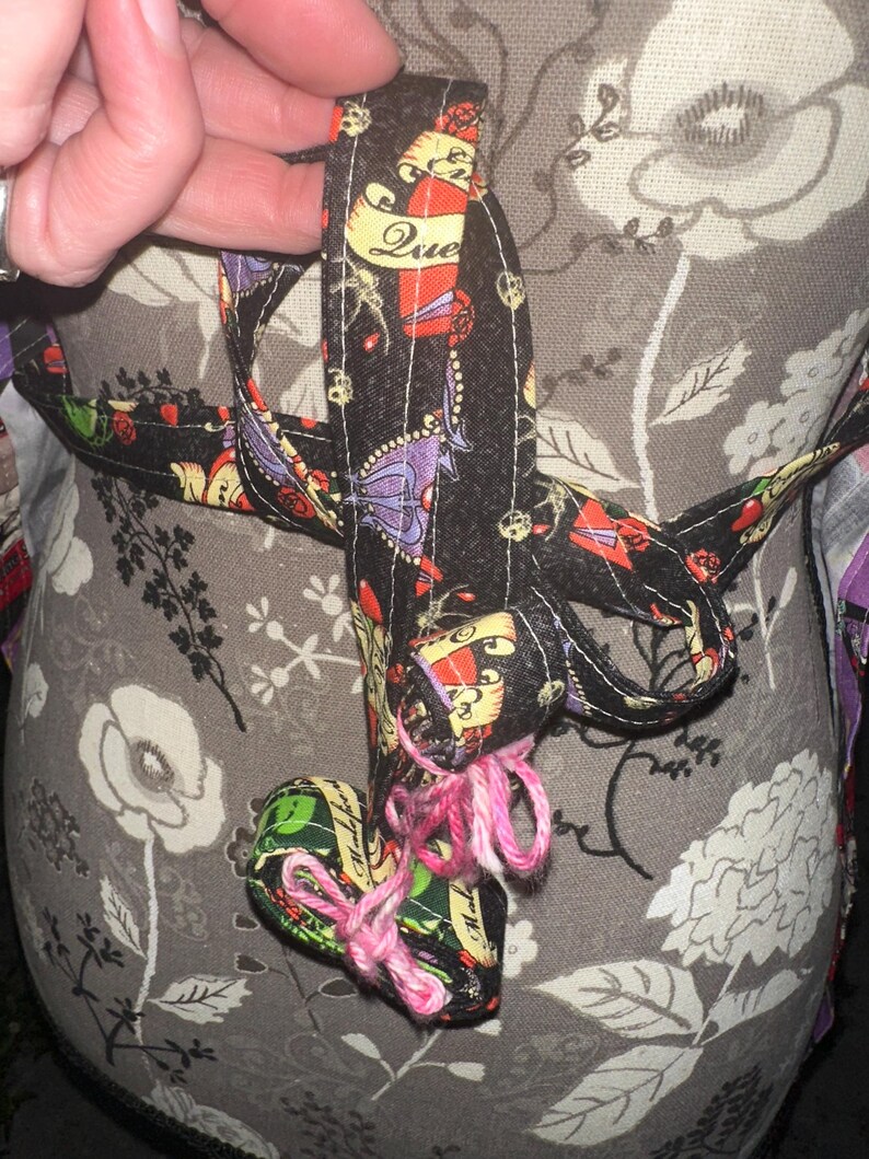 May include: A black fabric heart-shaped ornament with a pink yarn tie. The ornament is decorated with colorful tattoo-style designs and the word "Due". The background is a floral pattern.