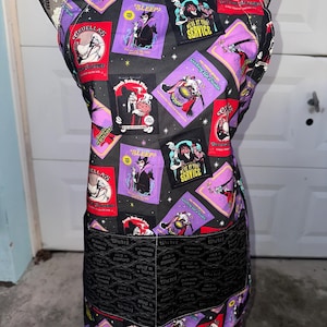 May include: Black apron with a repeating pattern of purple and red squares featuring cartoon villains. The apron has a black pocket with the word "Wicked" printed repeatedly. The apron has a black neck strap and ties.
