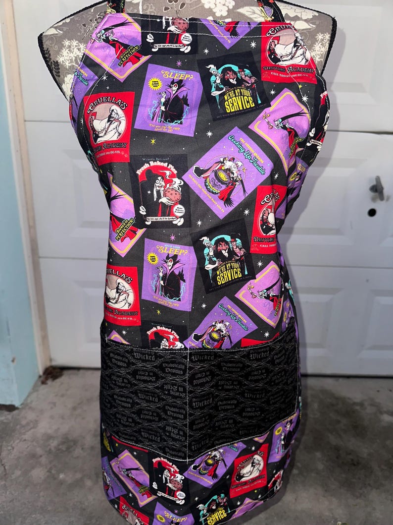 May include: Black apron with a repeating pattern of purple and red squares featuring cartoon villains. The apron has a black pocket with the word "Wicked" printed repeatedly. The apron has a black neck strap and ties.