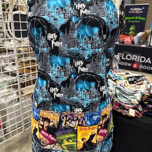 May include: A black and blue apron featuring a repeating pattern of Hogwarts and the words "Harry Potter." The apron has a pocket with images of the Harry Potter book covers. The apron is displayed on a mannequin.