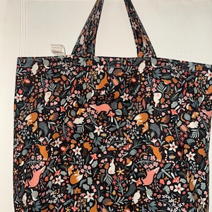 May include: A large tote bag with a black background and a colorful woodland pattern. The pattern includes foxes, rabbits, mushrooms, and flowers in shades of orange, pink, white, and green. The bag has long straps for carrying.