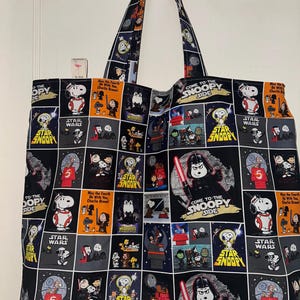 以下が含まれることがあります： スヌーピーとスターウォーズをテーマにしたコミックパネルの繰り返し模様が特徴の大きなトートバッグ。バッグは黒いハンドルと、オレンジ、グレー、白のアクセントが付いた黒い背景が特徴です。「Come to the Snoopy Side」と「Star Wars」の文字が含まれています。