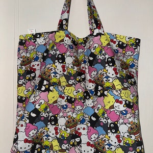 Puede incluir: Una bolsa de tela blanca con un estampado colorido de personajes de dibujos animados. Los personajes incluyen Hello Kitty, My Melody y Kuromi. La bolsa tiene dos asas negras.