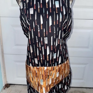 May include: A black apron with a repeating pattern of various knives in white, brown, and gray. The apron has a large pocket with a wood-grain design and a brown strap.