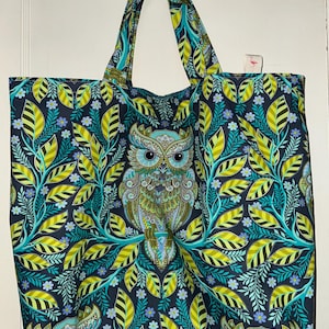 May include: A large tote bag featuring a detailed owl design. The bag's pattern includes teal, yellow, and green leaves and flowers against a dark background. The owl is centered, with intricate feather details.