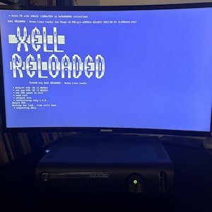 May include: A black Xbox 360 console sits beneath a Samsung monitor showing the text "XELL RELOADED" in white. The screen has a blue background with white text. The console's power button glows green.