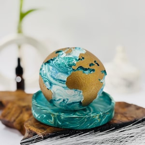 May include: A decorative globe with gold continents and swirling blue and white oceans sits atop a matching teal tray. The globe is a unique home decor item, perfect for a desk or shelf. The globe and tray are made of a marbled material.