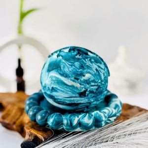 Może przedstawiać: Niebiesko-biała marmurowa kula spoczywa na pasującym pierścieniu z koralików. Kula i pierścień wykonane są z żywicy. Obiekty są prezentowane na drewnianej powierzchni z białym tłem.