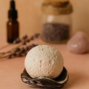 Puede incluir: Una esfera blanca texturizada, que se asemeja a la luna, descansa sobre un plato veteado en blanco y negro. El fondo borroso presenta una pequeña botella, un tarro y una piedra rosa. La imagen sugiere un tema de spa y autocuidado.