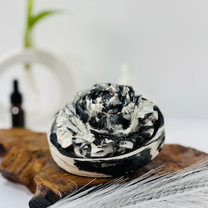 Op de afbeelding: Een decoratieve, ronde container met een gebeeldhouwd bloemmotief op het deksel. De container is zwart en wit, met een marmer effect. Het deksel is gevormd als een pioenroos. De container staat op een houten oppervlak. Diameter ca. 10 cm.