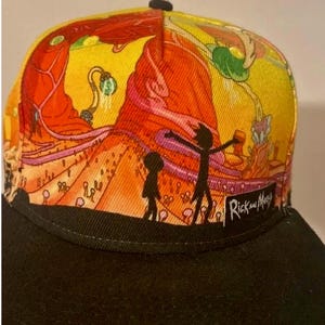 May include: A black and yellow baseball cap with a colorful graphic print of Rick and Morty characters. The cap has a black brim and a black strap with the words "Rick and Morty" printed on it.