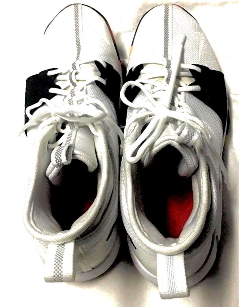 May include: A pair of white athletic shoes with black accents and white laces. The shoes have a mesh-like texture on the upper and a red interior. The soles are white with a gray stripe.