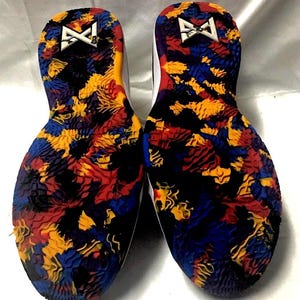 May include: Pair of athletic shoes with colorful soles. The soles feature a vibrant pattern of red, yellow, blue, and black shapes. The white midsoles have a red accent. The heel area displays a white logo.