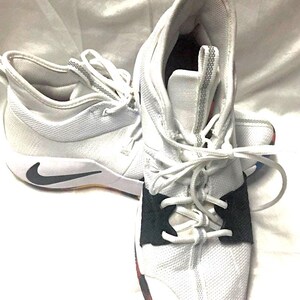 May include: White athletic shoes with black accents and a black Nike swoosh. The shoes have white laces and a black strap across the top of the foot. The soles are white with a black and red accent.