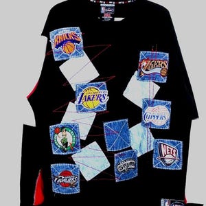 Op de afbeelding: Zwart langarm sweatshirt met verschillende NBA-teamlogo's die op de voorkant zijn genaaid. De logo's omvatten de New York Knicks, de Los Angeles Lakers, de Philadelphia 76ers, de Boston Celtics, de Cleveland Cavaliers, de San Antonio Spurs, de New Jersey Nets en het NBA-logo.