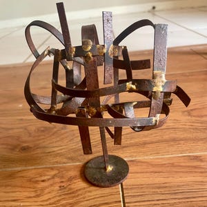 Puede incluir: Escultura abstracta de metal con un acabado marrón oxidado y detalles dorados. La escultura está hecha de finas tiras de metal que se entrelazan para crear una forma tridimensional. Está montada sobre una base redonda.