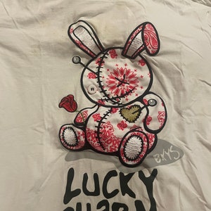 May include: White t-shirt with a red, white, and black embroidered design of a stitched bunny holding a red rose. The text "Lucky Charm" is printed in black below the bunny. The brand name "BKYS" is printed in red on the top of the shirt and in white below the bunny.