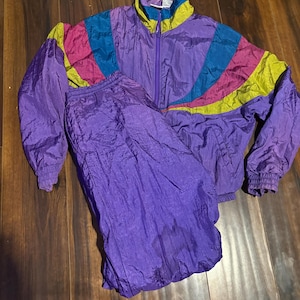 Op de afbeelding: Een vintage paarse trainingspakset. De jas heeft een ritssluiting aan de voorkant en een hoge kraag. De schouders hebben een colorblock-ontwerp met turquoise, roze en gele accenten. De bijpassende broek is effen paars. De set is gemaakt van glanzend, gekreukt materiaal.