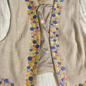 Pode incluir: Um colete de linho bege com uma guarnição decorativa de botões coloridos. Os botões estão dispostos em uma linha vertical de cada lado do colete. O colete tem uma etiqueta que diz "SQUARED RANCHWEAR".