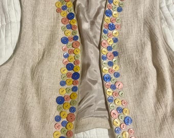 Puede incluir: Un chaleco de lino beige con un ribete decorativo de botones coloridos. Los botones están dispuestos en una línea vertical a cada lado del chaleco. El chaleco tiene una etiqueta que dice "SQUARED RANCHWEAR".