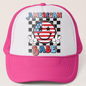 May include: A pink and white trucker hat with the words "AMERICAN BABE" in a retro font. The hat features a smiley face with the American flag design and peace signs. The background is a black and white checkered pattern.