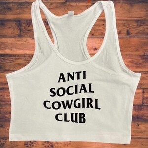 May include: White racerback tank top with the text "ANTI SOCIAL COWGIRL CLUB" in black. The tank top has a scoop neck and a racerback design. The background is a wooden surface.