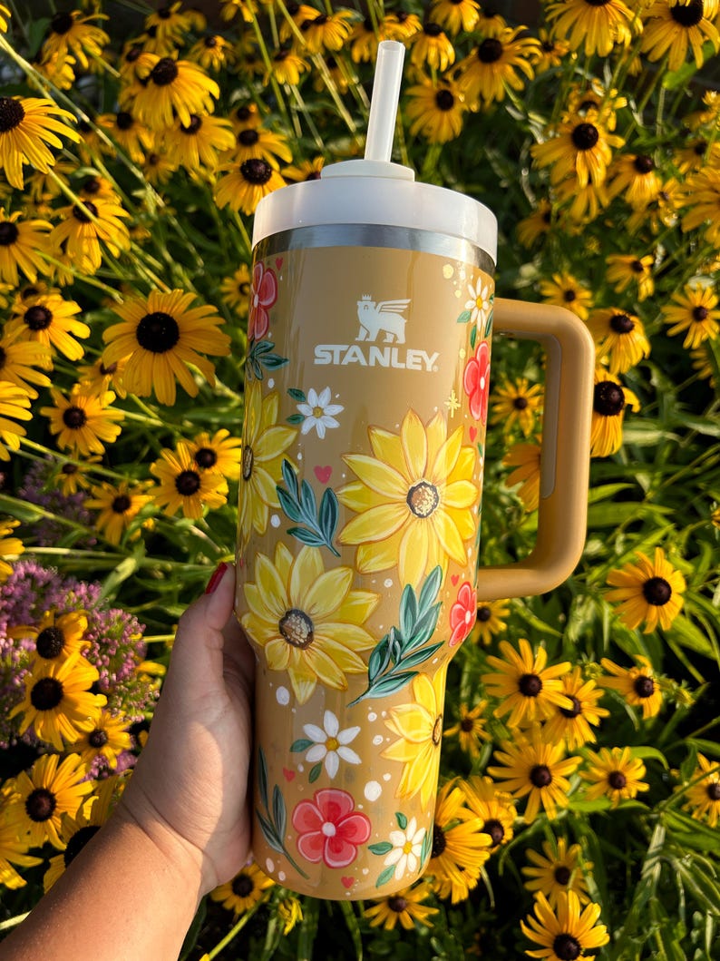 May include: A tan Stanley tumbler with a white lid and handle, adorned with a floral design featuring yellow sunflowers, red and white flowers, and green leaves. The tumbler has a white straw and the Stanley logo.