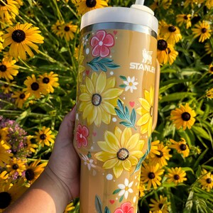 May include: A Stanley tumbler with a floral design featuring yellow sunflowers, pink and white flowers, and green leaves. The tumbler is tan with a white lid and straw. The background is a field of yellow flowers.